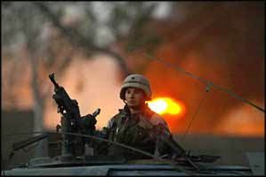 A USA occupier guards the burning remains of a factory in Baghdad, which was hit by Iraqi Resistance mortar fire.